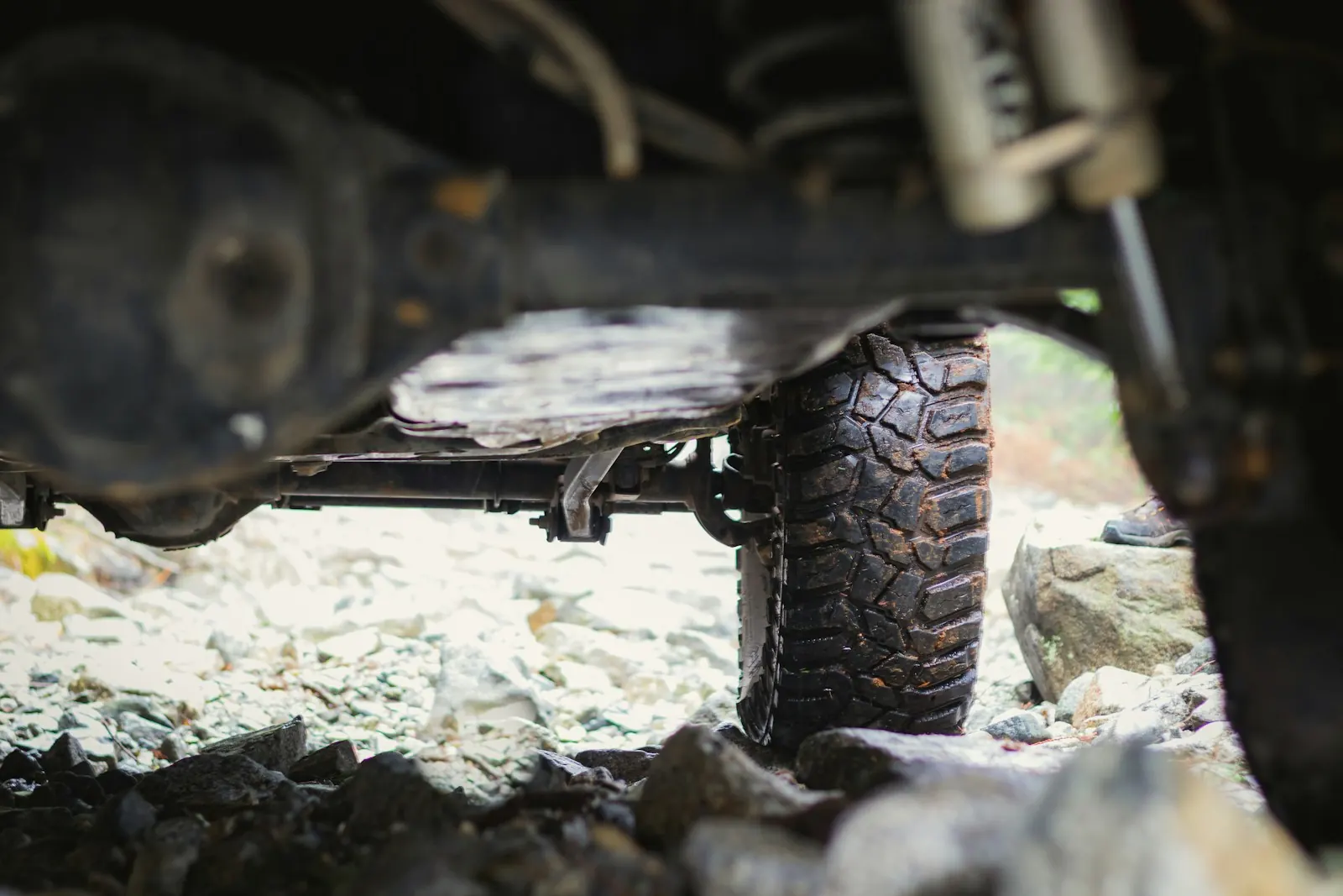 Low view under a 4x4 of a solid axle housing and muddy knobby tires on wet rocks