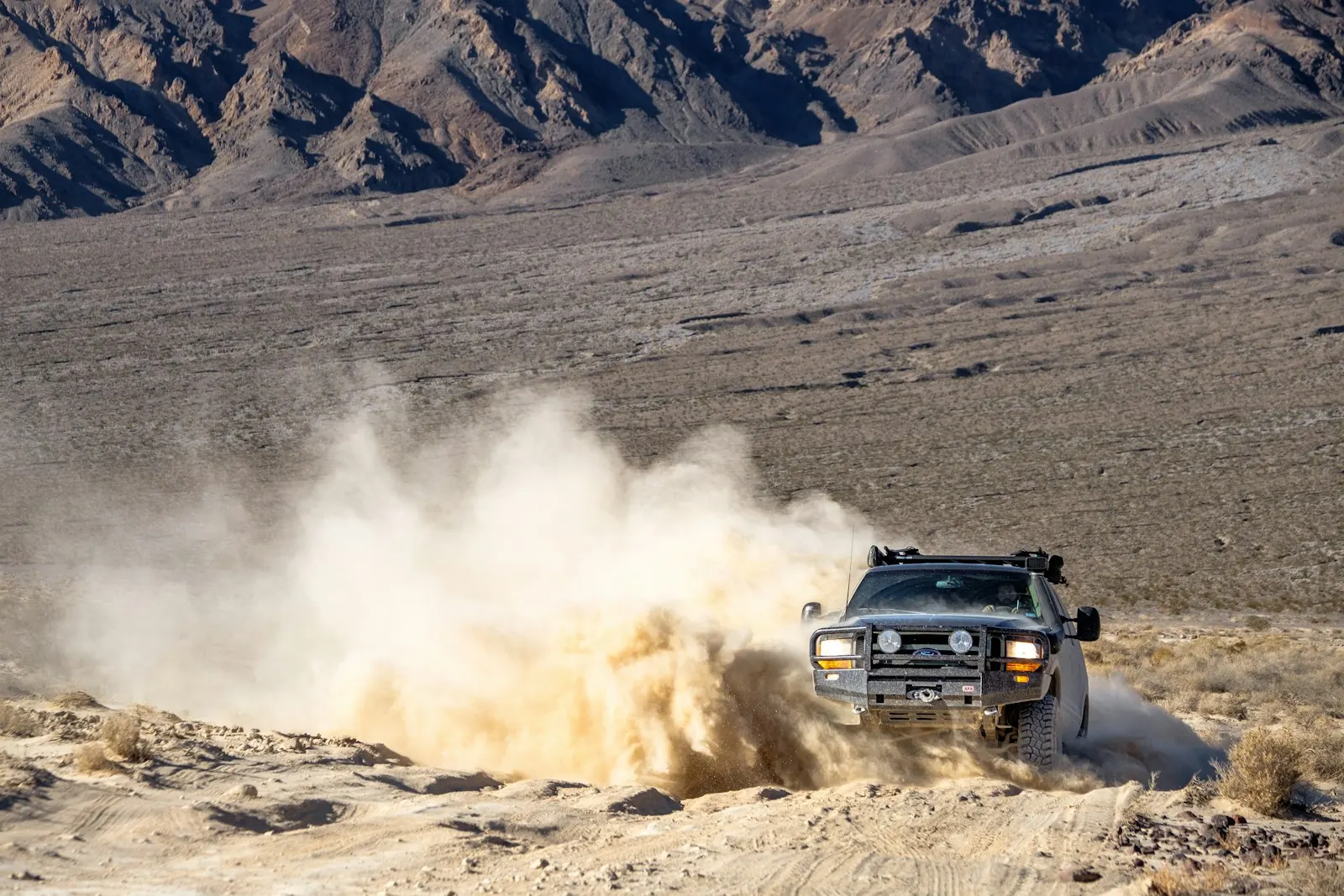 Built pickup with a steel bumper, lights and roof rack kicking up dust on a desert trail below mountains