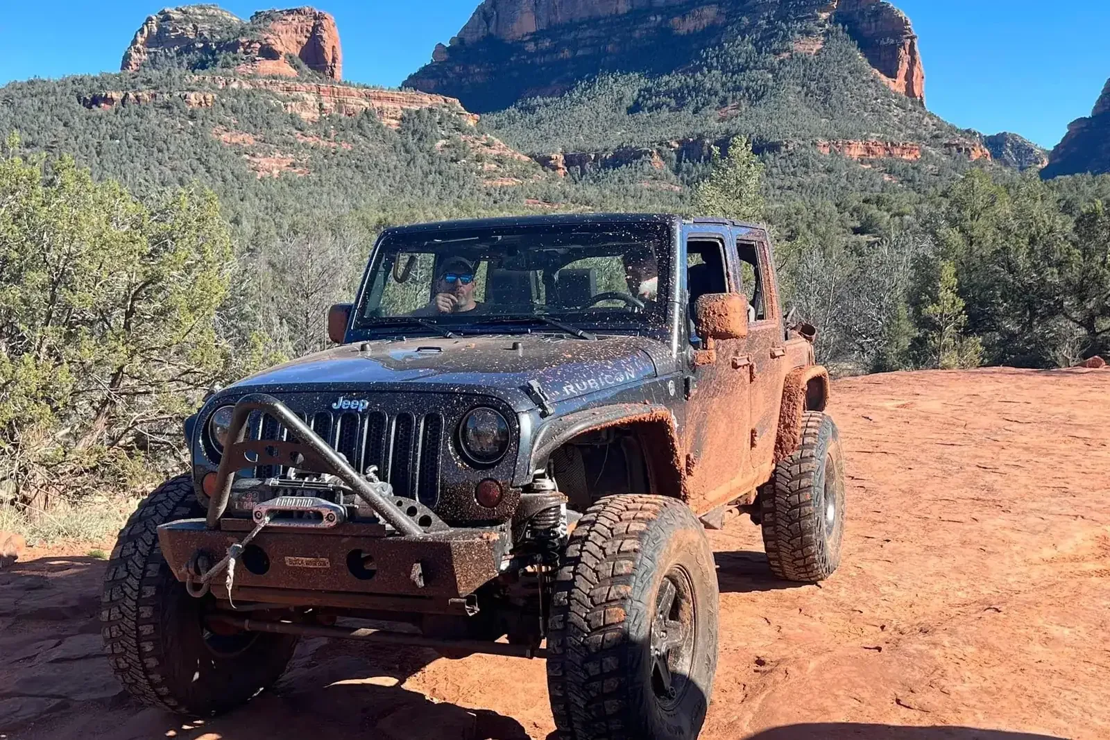 Four-door Wrangler with a winch bumper parked on red dirt below Sedona rock formations